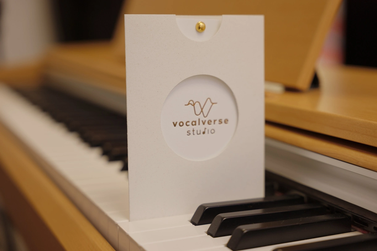 Zdjęcie Vouchera - białe zaproszenie z wycięciem w kształcie koła, gdzie znajduje się napis Vocalverse Studio. Karka stoi na pianinie.